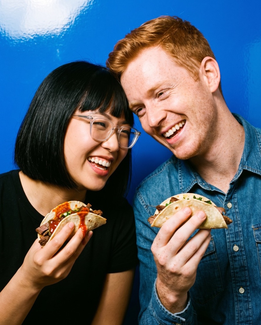 Hyeri Bang and Callum Pryce leaning together and laughing over street tacos loaded with red chili sauce.