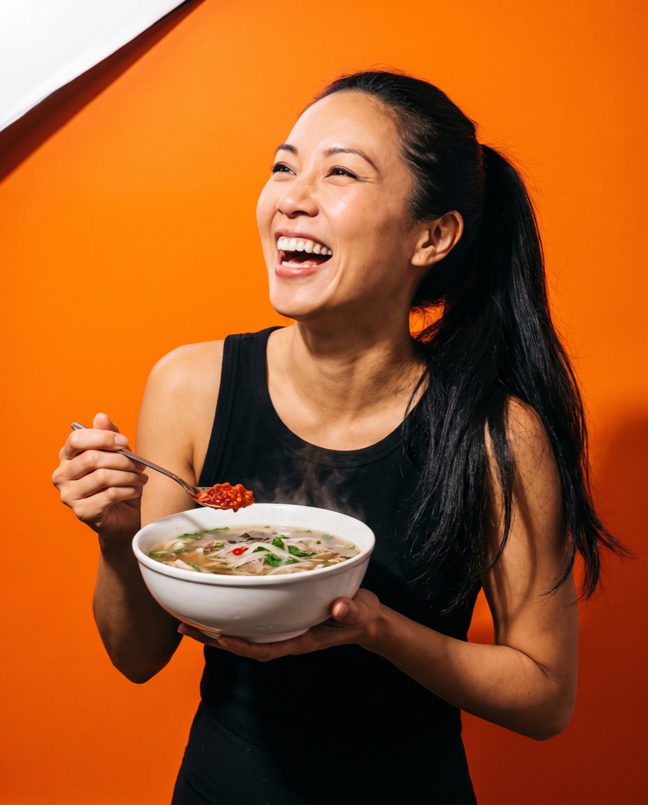 Linh Tran laughing with her head tipped back, holding a bowl of pho with a spoon of sambal over the broth.