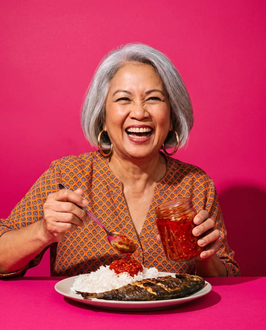 Remedios Bacani laughing while spooning sambal onto a plate of rice and grilled fish.