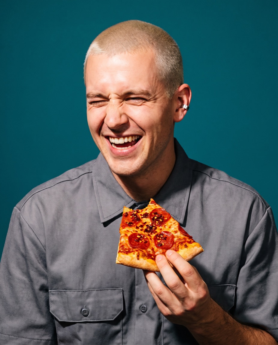 Wes Kolditz wincing and laughing while eating a folded slice of pizza covered in chili crisp.