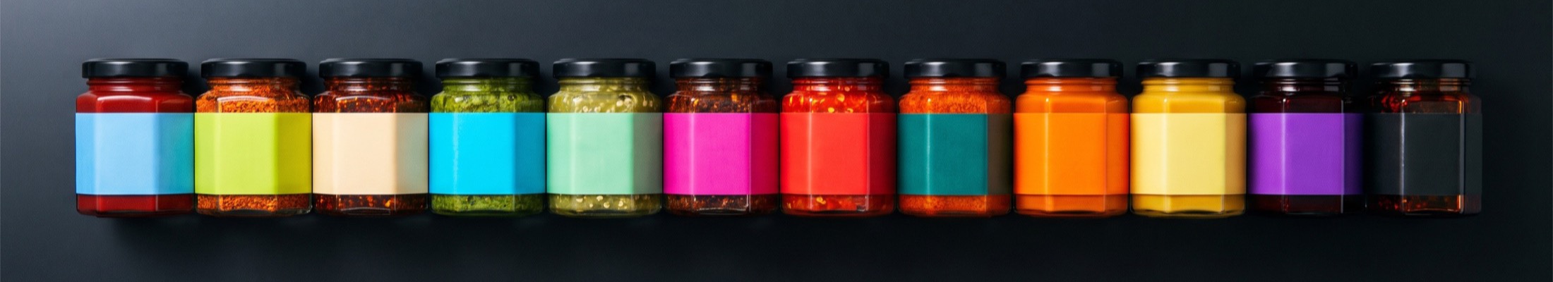 Twelve jars of sambal photographed from overhead in a row. Each holds a visibly different paste, from pure red through greens to near black, under a differently coloured label band.