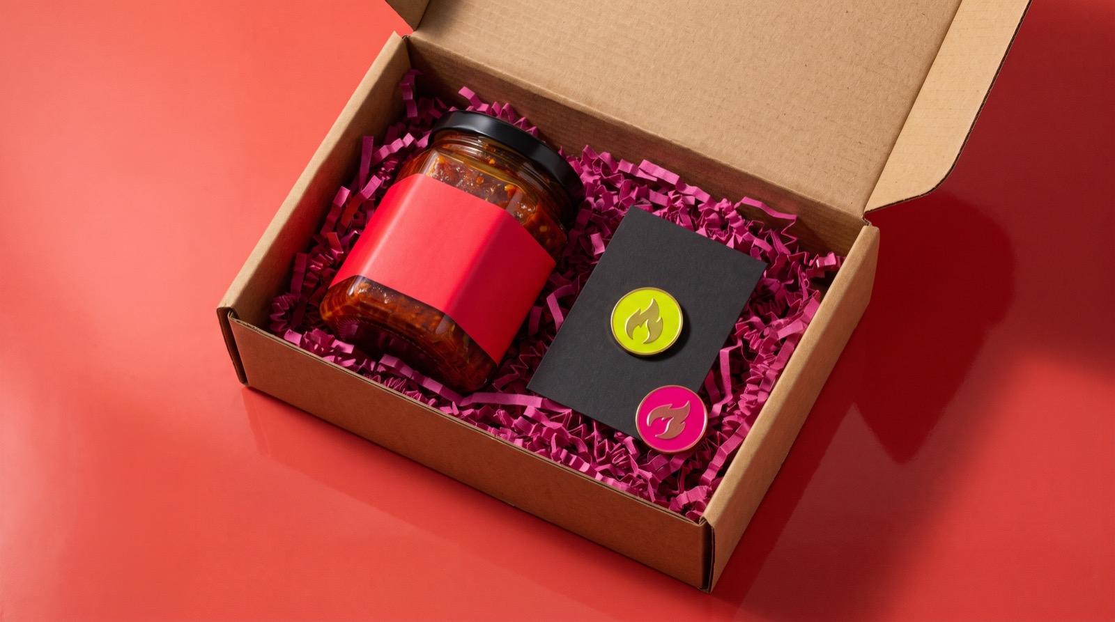 An open shipping box holding a jar of sambal and a black card with two enamel pin badges attached.
